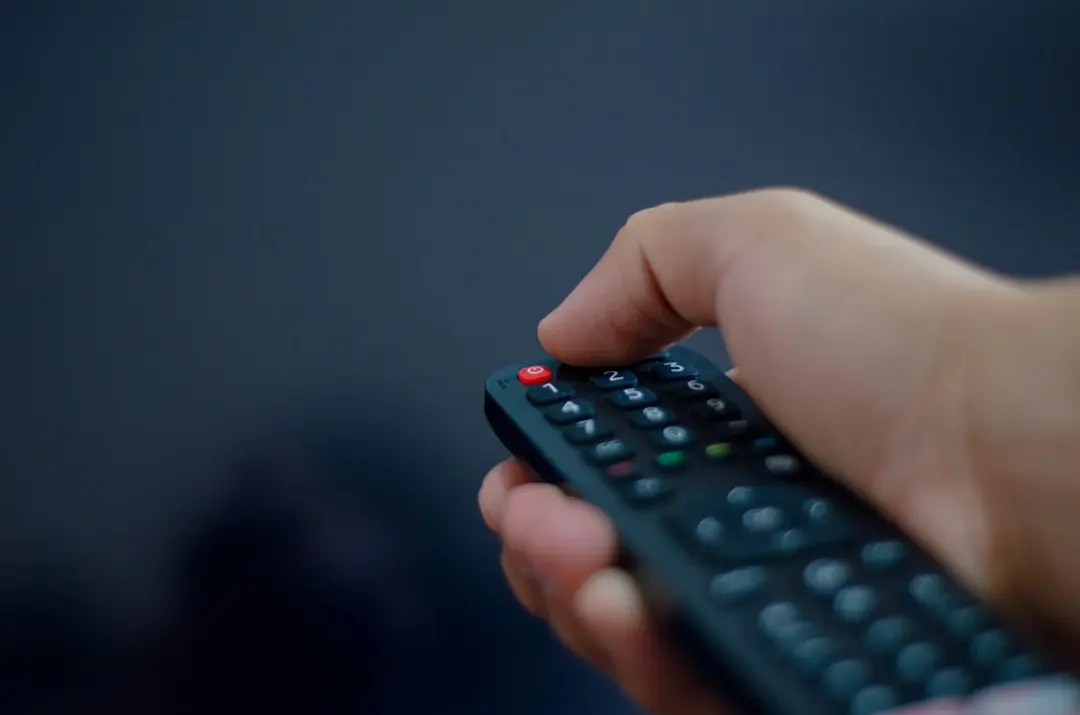 A hand pressing the buttons of a black TV remote control against a dark blue background