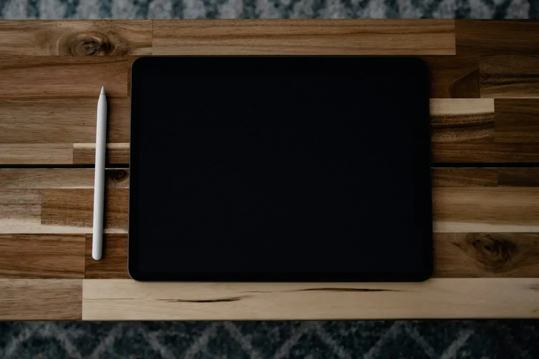 A tablet with a dark screen and a white stylus lying on a wooden table, seen from above